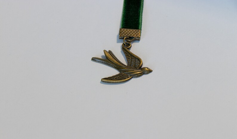 May include: A green velvet bookmark with a metal bird charm. The bird is in flight and has a detailed design.