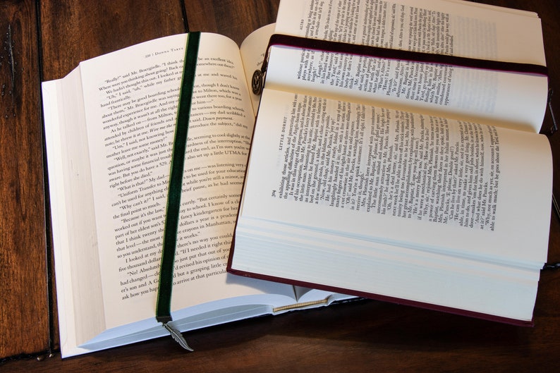 May include: Two open books with bookmarks. The book on the left has a green velvet bookmark with a feather charm. The book on the right has a burgundy velvet bookmark.
