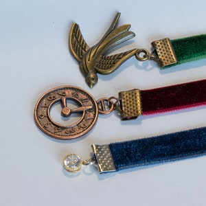 May include: Three velvet ribbon bookmarks with decorative metal charms. The charms include a bird, a clock face, and a rhinestone.
