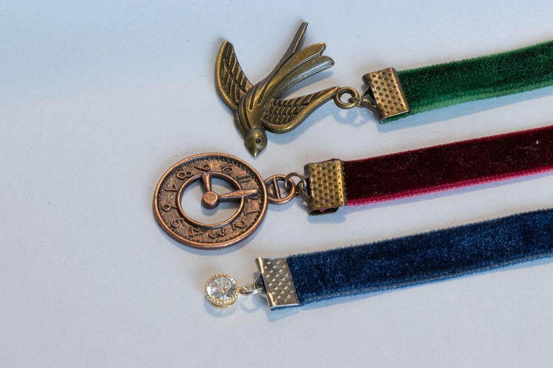 May include: Three velvet ribbon bookmarks with decorative metal charms. The charms include a bird, a clock face, and a rhinestone.