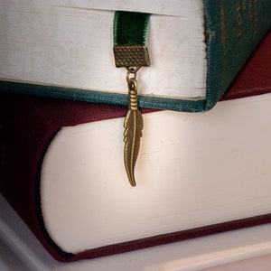 May include: Two books stacked on top of each other. The top book has a green cover and a velvet bookmark with a feather charm. The bottom book has a burgundy cover.