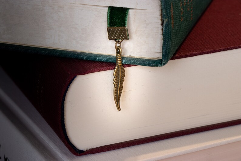 May include: Two books stacked on top of each other. The top book has a green cover and a velvet bookmark with a feather charm. The bottom book has a burgundy cover.