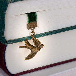 May include: A green and burgundy book with a green velvet bookmark. The bookmark has a gold metal charm shaped like a bird.