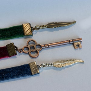 May include: Three velvet ribbon bookmarks with different colored ribbons and decorative metal charms. The charms include a feather, a key, and another feather.