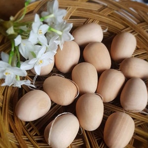 May include: A collection of light brown wooden eggs with twine loops, nestled in a woven basket. White flowers with yellow centers and green stems are arranged on the left side of the basket. The eggs have a smooth, natural finish.