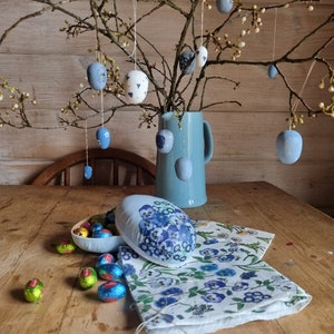 May include: An Easter-themed still life featuring hanging painted eggs, a blue pitcher, and a floral-patterned napkin. The eggs are in various shades of blue and white, some with floral designs. Chocolate eggs in colorful foil are also present.