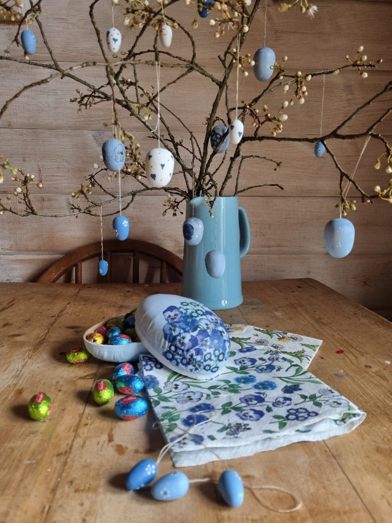May include: An Easter-themed still life featuring hanging painted eggs, a blue pitcher, and a floral-patterned napkin. The eggs are in various shades of blue and white, some with floral designs. Chocolate eggs in colorful foil are also present.