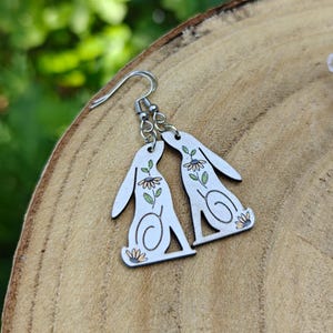 Puede incluir: Pendientes plateados con diseño de conejito. Los pendientes son blancos con un diseño floral en verde, amarillo y negro. Los pendientes cuelgan sobre una superficie de madera.