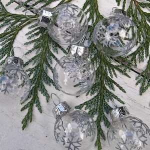 Puede incluir: Cinco adornos esféricos transparentes con diseños de copos de nieve con purpurina plateada. Cada adorno tiene una tapa de metal plateado con un pequeño lazo para colgar. Los adornos están dispuestos en un grupo y son transparentes.