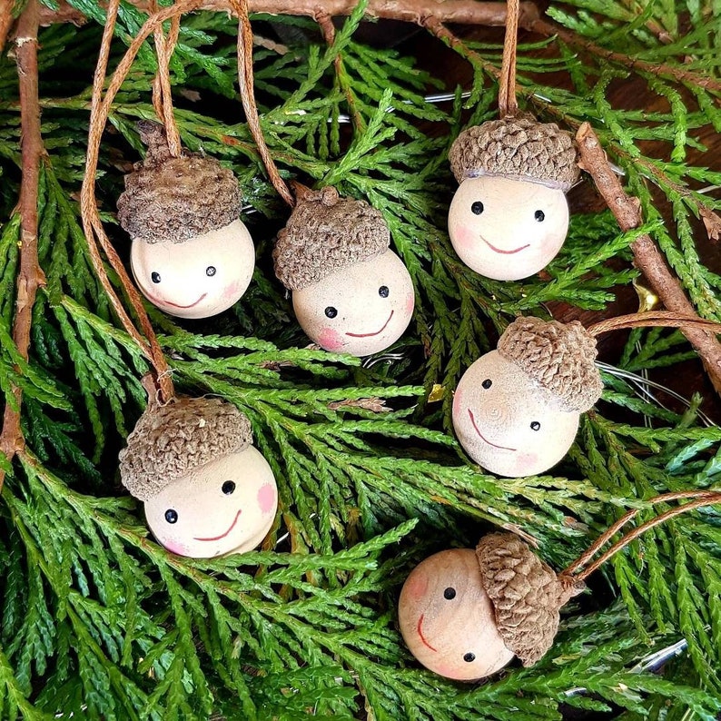 May include: Six brown acorn ornaments with painted faces and smiles. The acorns are hanging from twine and are surrounded by green pine branches.