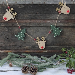 Puede incluir: Una guirnalda decorativa con tres cabezas de reno de madera clara con astas de purpurina dorada y narices de pompón rojo, intercaladas con dos árboles de Navidad de madera verde oscuro con lunares blancos, unidos por un cordel marrón.