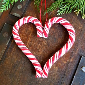 Puede incluir: Adorno navide&ntilde;o en forma de coraz&oacute;n hecho con dos bastones de caramelo. Los bastones tienen rayas rojas y blancas, con una cinta roja para colgar. El adorno est&aacute; sobre un fondo de madera r&uacute;stica con vegetaci&oacute;n.