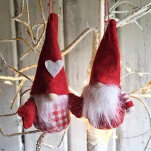 May include: Two hanging gnome decorations. Each gnome has a red pointed hat, a white beard, and a small nose. One gnome has a white heart on its hat and a red and white checked body. The other gnome has a striped arm.