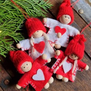 May include: Four red felt Christmas decorations shaped like people with white felt hearts on their chests. Each decoration has a white felt face with a painted smile and black eyes. The decorations are hanging from a green pine branch.