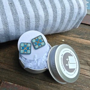 May include: A pair of square stud earrings featuring a light blue and gold geometric star pattern. The earrings are displayed on a white circular card inside a small round metal tin with a lid that reads handmade by www.hyggestyle.co.uk.