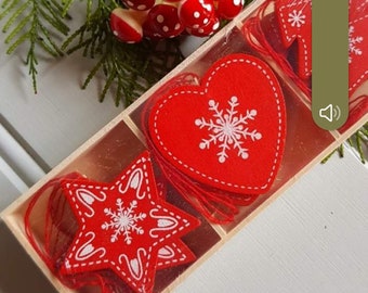 Puede incluir: Un conjunto de adornos de fieltro rojo en una caja de madera rectangular, que incluye una forma de corazón con un estampado de copo de nieve blanco y una forma de estrella con patrones decorativos blancos. Cada adorno tiene un cordel rojo.