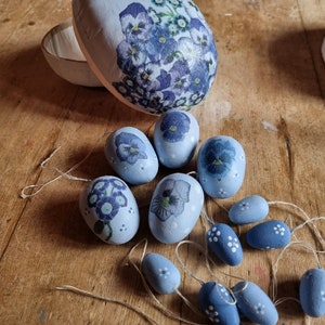 May include: A collection of light blue decorative Easter eggs and ornaments. The eggs are adorned with floral designs in shades of blue and purple. Some eggs are open, revealing a white interior. The ornaments are strung on twine.