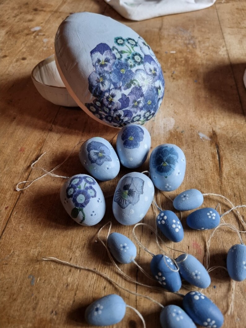 May include: A collection of light blue decorative Easter eggs and ornaments. The eggs are adorned with floral designs in shades of blue and purple. Some eggs are open, revealing a white interior. The ornaments are strung on twine.