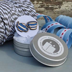 May include: A pair of wooden earrings shaped like a teapot and a teacup. The earrings are painted blue and white with a striped pattern. The earrings are packaged in a small silver tin with a label that reads "handmade by www.hyggestyle.co.uk".