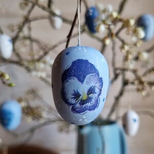 May include: A light blue Easter egg with a hand-painted purple pansy flower design. The egg is suspended by a string, with other decorated eggs and branches in the background. The egg has white polka dots.