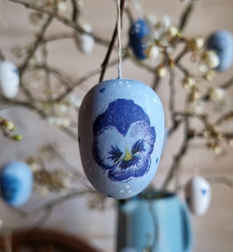 May include: A light blue Easter egg with a hand-painted purple pansy flower design. The egg is suspended by a string, with other decorated eggs and branches in the background. The egg has white polka dots.
