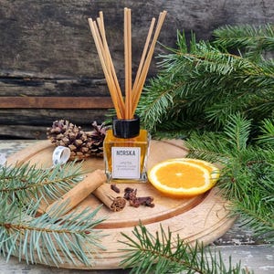 May include: A glass bottle diffuser with wooden sticks, filled with a golden liquid. The bottle has a black label with the text "Norska" and "Vinter Orange, Clove & Cinnamon". The diffuser is surrounded by pine branches, cinnamon sticks, cloves, and an orange slice.