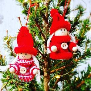 Puede incluir: Dos adornos colgantes con forma de pequeñas figuras con cabezas de madera, caras pintadas y pelo de lana blanca. Cada una lleva un gorro de punto rojo brillante y un suéter a juego con un botón blanco en la parte delantera.
