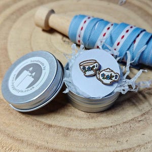 May include: A small tin with a white background and blue floral teacup and teapot designs. The tin has a silver lid with a sticker that says "handmade by www.hyggestyle.co.uk".