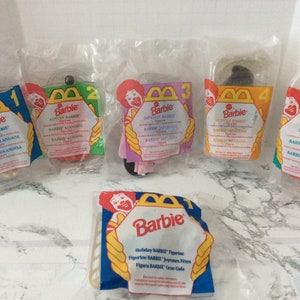 May include: Six different McDonald's Happy Meal toys featuring Barbie dolls from different countries. The toys are packaged in clear plastic bags with colorful labels. The labels feature the McDonald's logo and the Barbie logo. The text on the labels includes the names of the countries represented by the dolls, such as "Dutch Barbie", "Japanese Barbie", and "USA Barbie".
