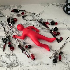May include: A set of six phone charms featuring various bloody weapons, including a knife, scissors, an axe, and a butcher knife. Each charm is adorned with black and red beads.