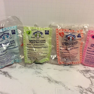 May include: Four plastic packages with Cabbage Patch Kids toys inside. Each package has a different color and a number from 1 to 4. The packages have a Happy Meal logo and text that says "Congratulations on your new arrival!" with a name and the hospital where the baby was born.