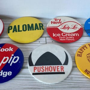 May include: Seven round, colorful buttons with various designs and text. The buttons feature a heart with the text "I GWMC", a yellow button with the text "PALOMAR", a button with the text "Try New Lady Lee Ice Cream Finer Quality New Flavors", a blue button with the text "The New MAIBasic Four", a button with the text "I Took The pip Pledge", a white button with the text "PUSHOVER", and a yellow button with the text "HAPPY NEW YEAR! NOVEMBER 20, 1982".