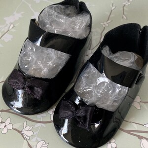 May include: A pair of black patent leather baby shoes with black satin bows. The shoes have a strap across the top and a buckle closure.