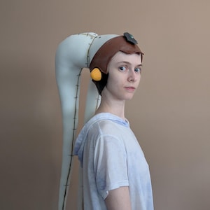 Könnte beinhalten: Ein weißer Twi'lek-Kopfschmuck mit zwei langen, gepolsterten Lekku, die entlang der Nähte dunkle Steppungen aufweisen. Der Kopfschmuck umfasst ein braunes, lederartiges Stirnband, ein dunkles Akzentstück an der Stirn und gelbe Ohrabdeckungen.
