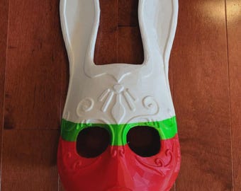 May include: A white, red, and green bunny mask with large ears. The mask has a decorative design on the forehead and around the eyes. The lower half of the mask is red, with a green stripe separating it from the white upper half.