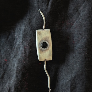 Könnte beinhalten: Ein weißer rechteckiger Anhänger mit einem schwarzen runden Stein in der Mitte. Der Anhänger ist an einer weißen Schnur befestigt.