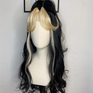 May include: A long, black wig with blonde streaks. The wig is styled in loose waves and has a side parting. The wig is on a mannequin head.