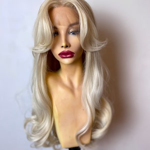 May include: A long, wavy, platinum blonde wig with a lace front. The wig is styled with loose waves and a side part. The wig is on a mannequin head.