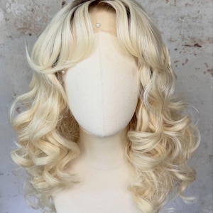 May include: A blonde wig with dark roots and soft, voluminous curls. The wig is styled with a side part and features a natural-looking hairline. The wig is displayed on a mannequin head.