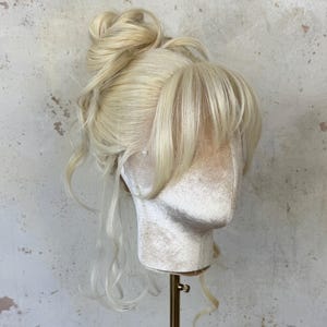 Op de afbeelding: Een platinablonde pruik gestyled in een hoge knot met losse lokken en een lange, zwierige pony. Het haar is in een opgestoken kapsel met een zachte, golvende textuur geplaatst op een mannequin-hoofd in een neutrale kleur.