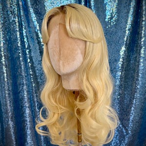 Puede incluir: Una peluca rubia con cabello largo y ondulado y un flequillo lateral se muestra en una cabeza de maniquí. La peluca tiene una línea de cabello de aspecto natural y está peinada con rizos sueltos. El fondo es una tela azul brillante.
