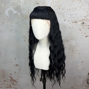 May include: A black wig with long, wavy hair and a blunt fringe, styled on a mannequin head. The wig features a braided detail at the crown and is displayed against a mottled, neutral-toned background.