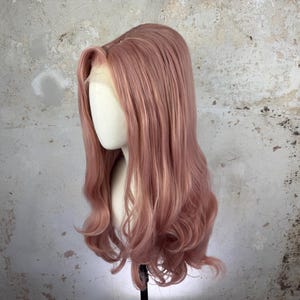 Könnte beinhalten: Eine lange, wellige Perücke in Staubrosa mit dezenten blonden Highlights. Die Perücke ist mit einem Seitenscheitel und einer Lace-Front-Konstruktion versehen und wird auf einem Mannequinkopf präsentiert. Das Haar fällt in weichen Locken.