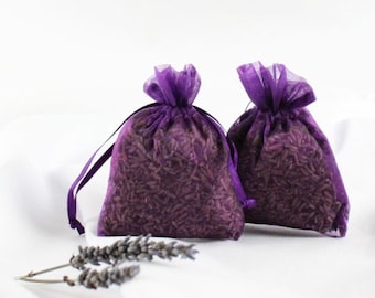 May include: Two small, semi-transparent purple organza drawstring bags filled with dark purple grains. The bags are cinched at the top with matching purple ribbons. Two dried lavender sprigs with greyish-purple buds lie in front.