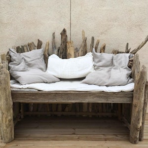 May include: A rustic wooden bench made from driftwood with a white linen cushion and two gray linen pillows. The bench is sitting on a wooden deck.