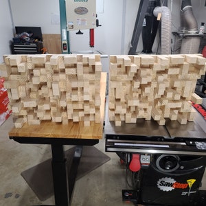 May include: Two wooden acoustic panels made of small, rectangular blocks of wood. The panels are sitting on a workbench with a black and silver table saw in the background.