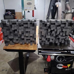May include: Two gray acoustic foam panels with a 3D cube design. The panels are displayed on a wooden table and a black table saw. The panels are designed to absorb sound and reduce echo in a room.