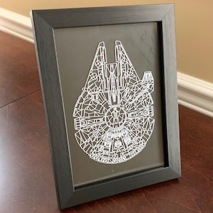 May include: A black framed picture with a white geometric design of the Millennium Falcon spaceship from Star Wars.