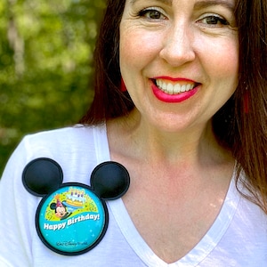 May include: A black Mickey Mouse ear button with a round Happy Birthday! design. The button features a cartoon image of Mickey Mouse and a birthday cake. The text "Happy Birthday!" is in white on a blue background.