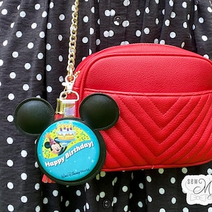 May include: A red crossbody bag with a chevron pattern and a gold chain strap. A Mickey Mouse-shaped keychain with a "Happy Birthday!" design is attached. The bag is shown against a black and white polka dot dress.
